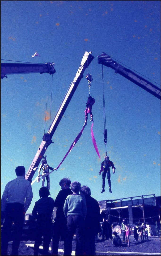 The image shows a blue-tinted color photograph taken during the performance or rehearsal of Leopoldo Maler’s Crane Ballet. In the foreground, several figures stand with their backs to the camera, looking at three performers suspended in the air by harnesses attached to the individual arms of three mobile cranes. The performers hang loosely, connected to one another by thick bands of red ribbon trailing down from the harness of the central and highest performer to the two others on either side of the image below him.