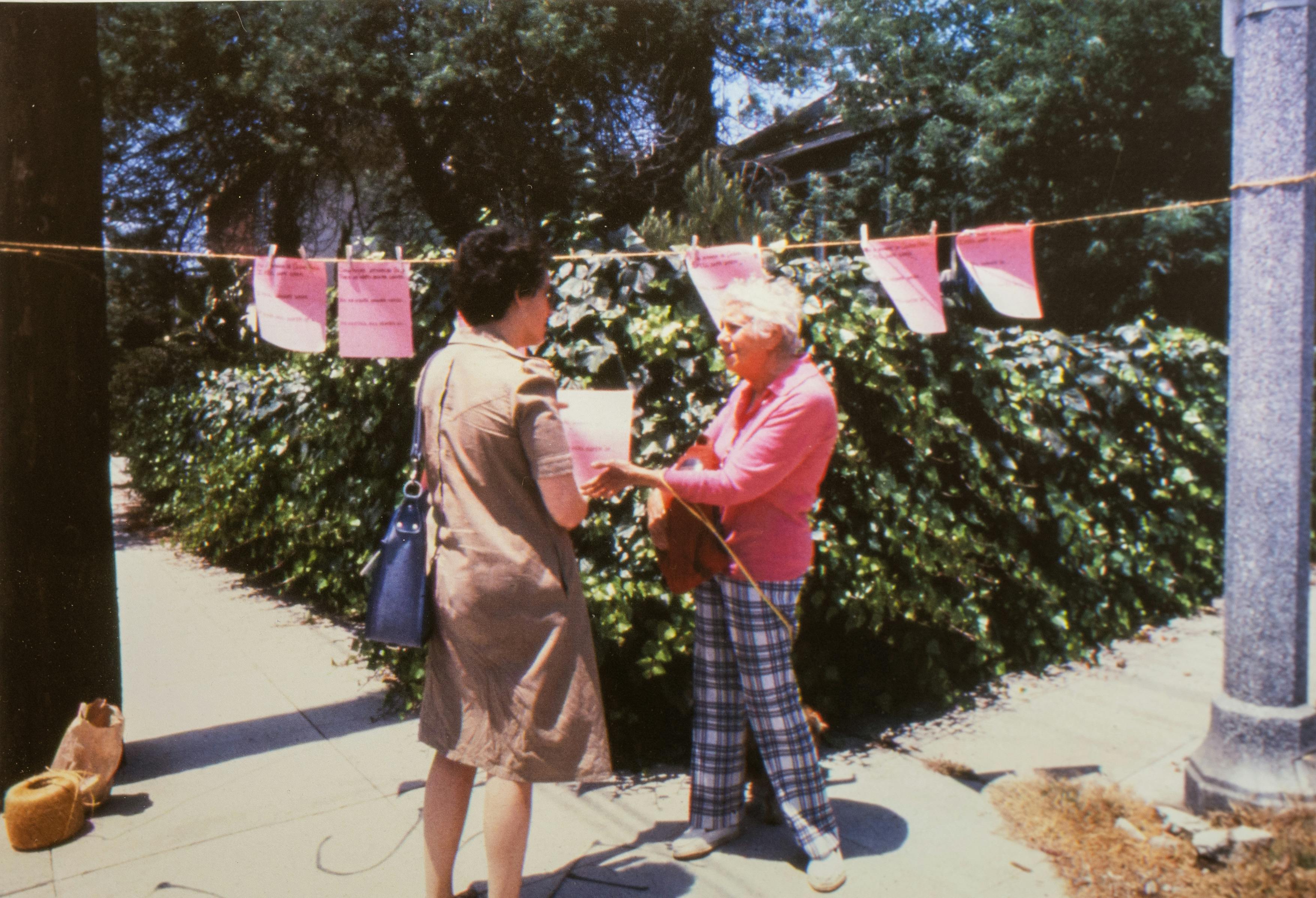 A color photograph, at the center of which two women are speaking. One woman, who has her back to the camera, hands the other a pink flyer, with words that are not legible in the photograph. They stand between a telephone pole, on the left, and a streetlamp, on the right, from which a clothesline has been hung. Clipped to the clothesline are more pink sheets of paper with text.