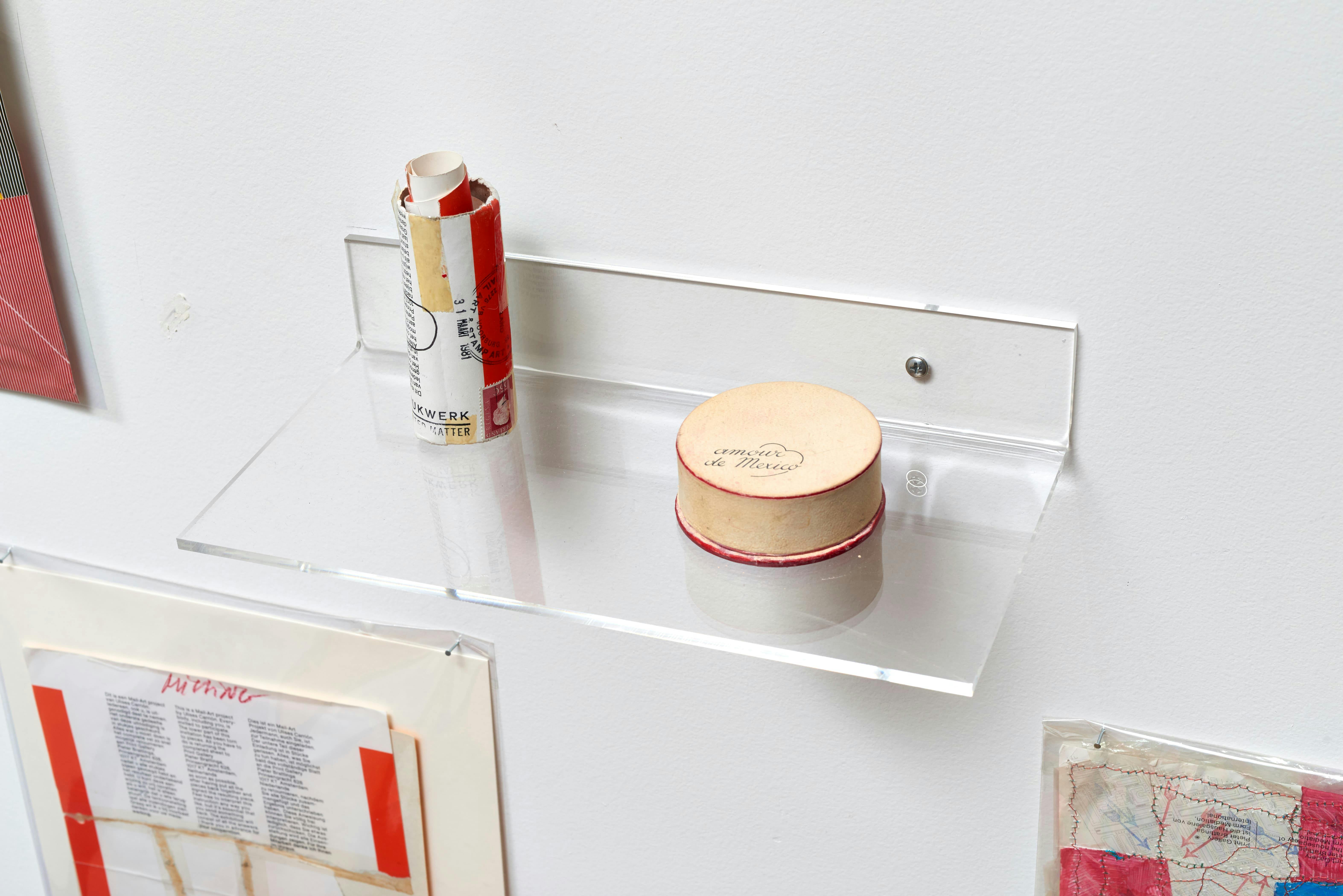 Close up view of a powder compact and rolled up paper positioned on a clear, wall-mounted shelf.