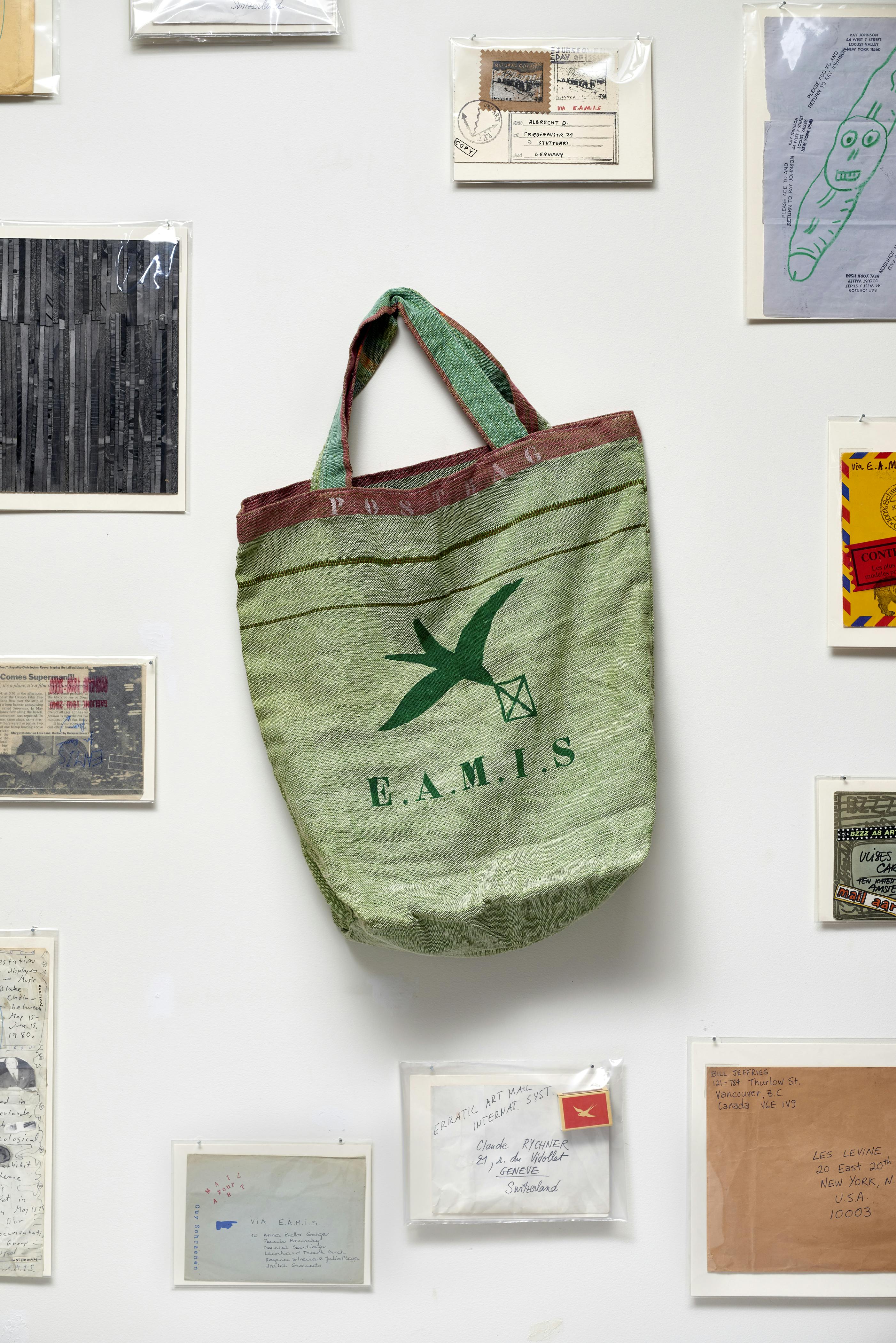 Installation view of Ulises Carrión exhibition showing a green tote bag mounted to a wall surrounded by photographs, sketches, and envelopes.
