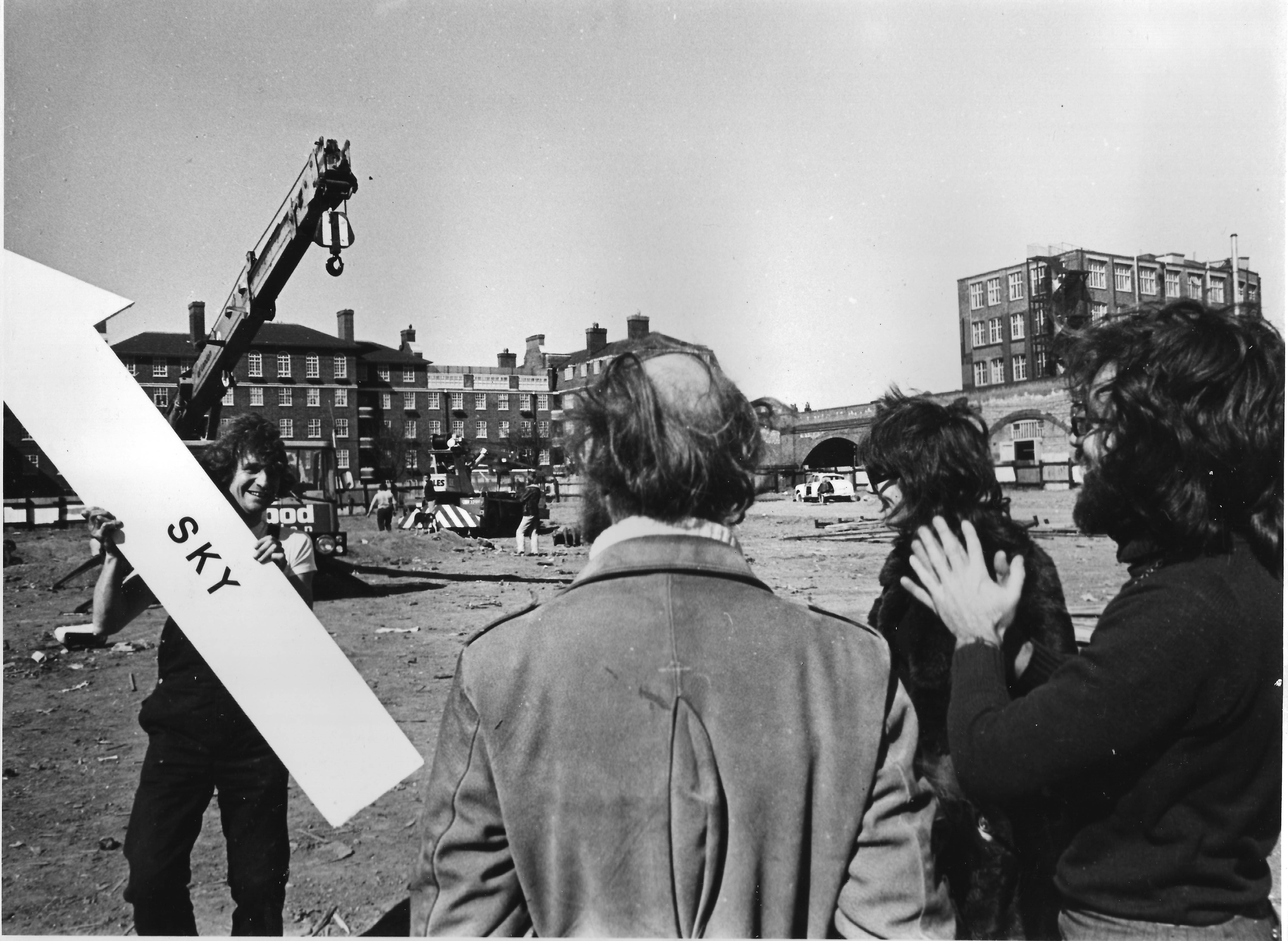 The image shows a black-and-white photograph taken during the performance or rehearsal of Crane Ballet. In the foreground, three figures stand with their backs to the camera, looking out at an unpaved construction site where two mobile cranes are visible. In the image’s left foreground, a young man holding a large white arrow with the word “SKY” written on it, and pointing upward, appears to be showing the sign to the three figures. The figure on the far right, the artist Leopoldo Maler, claps his hands while looking at the man holding the sign.