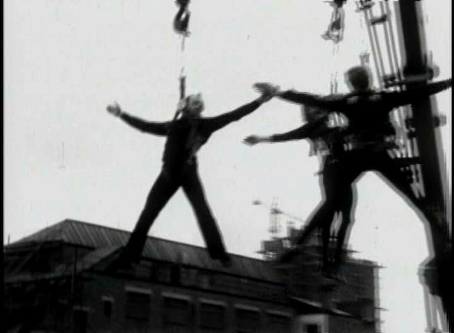 The image is a still from a black-and-white video documenting the performance of Leopoldo Maler’s Crane Ballet for the BBC. In the image, three acrobats hang suspended in the air, with their arms and legs outstretched in rough imitations of the letter “X.” Each performer is harnessed to the arm of a respective crane.