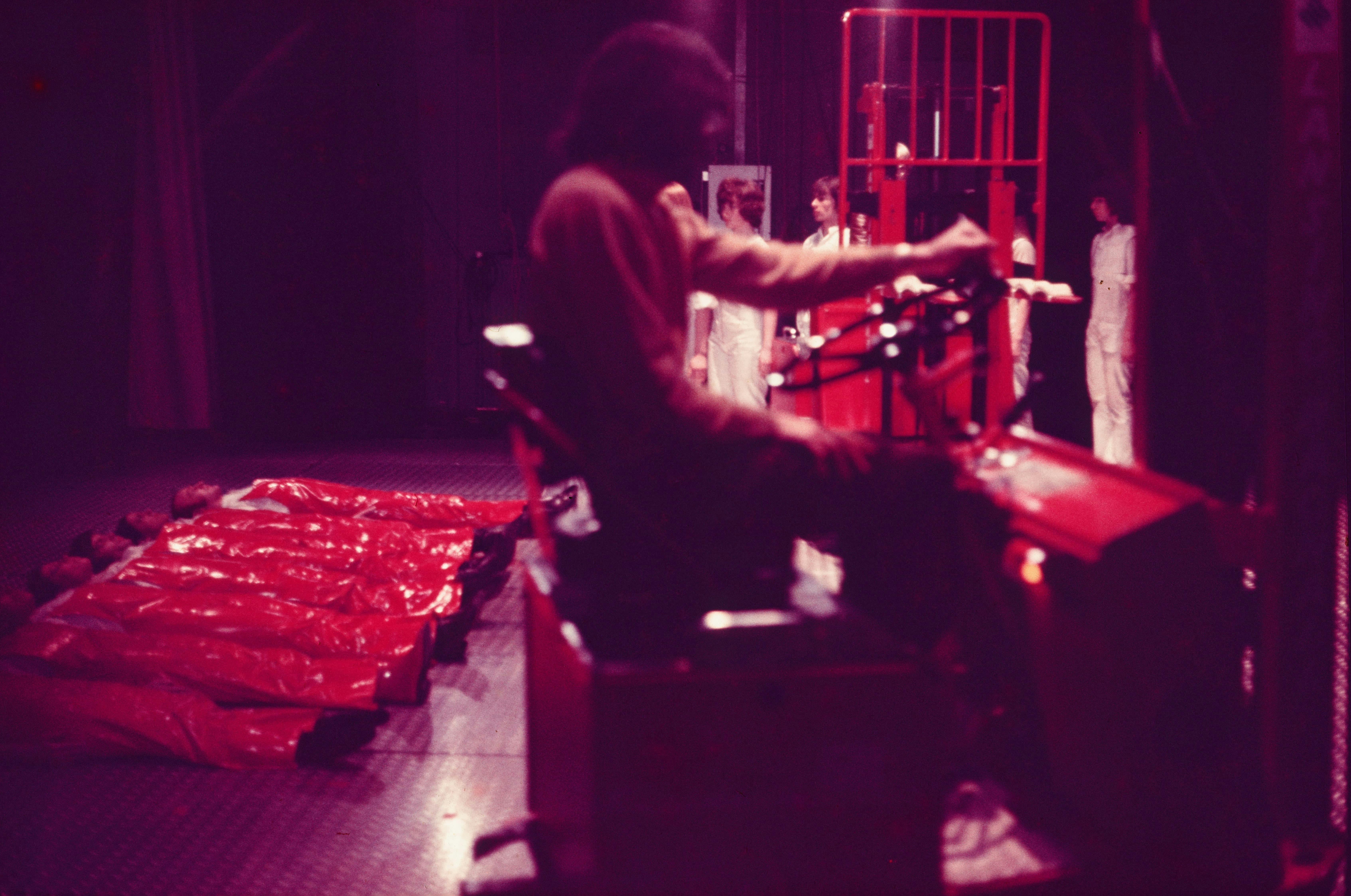 A color photograph. In the foreground, taking up most of the right side of the image, a man appears mid-motion driving a red forklift. To his left, performers wearing red vinyl costumes lie face up in a line on an industrial floor, their arms above their heads and their legs kicked up against a corresponding line of standing performers in white, who are holding their feet. At the feet of this group, and behind the driven forklift, several standing figures in white approach the line of performers in red in a loose formation. 