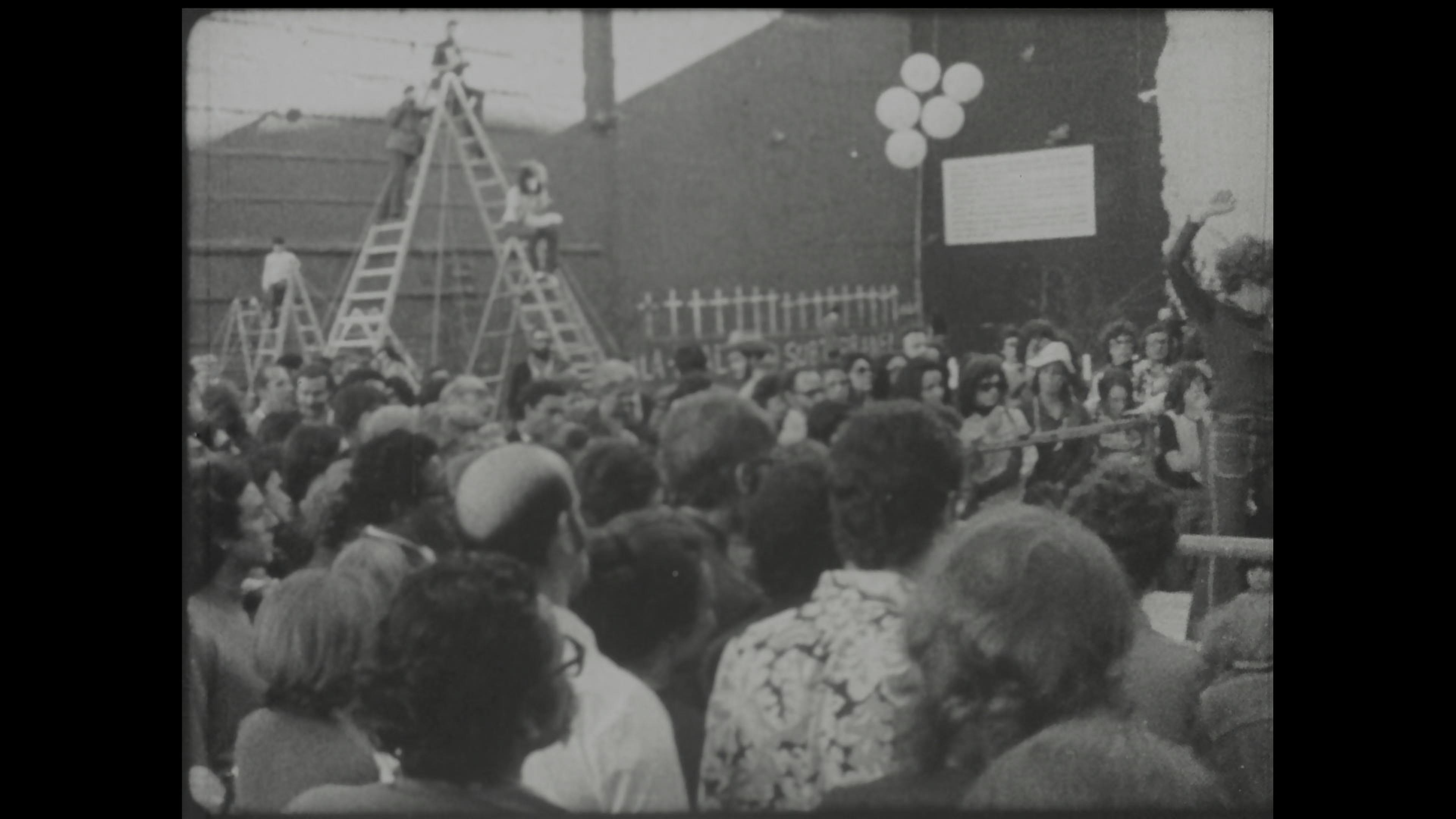 Black-and-white film still of crowd gathered in a public space with a ladder in the background