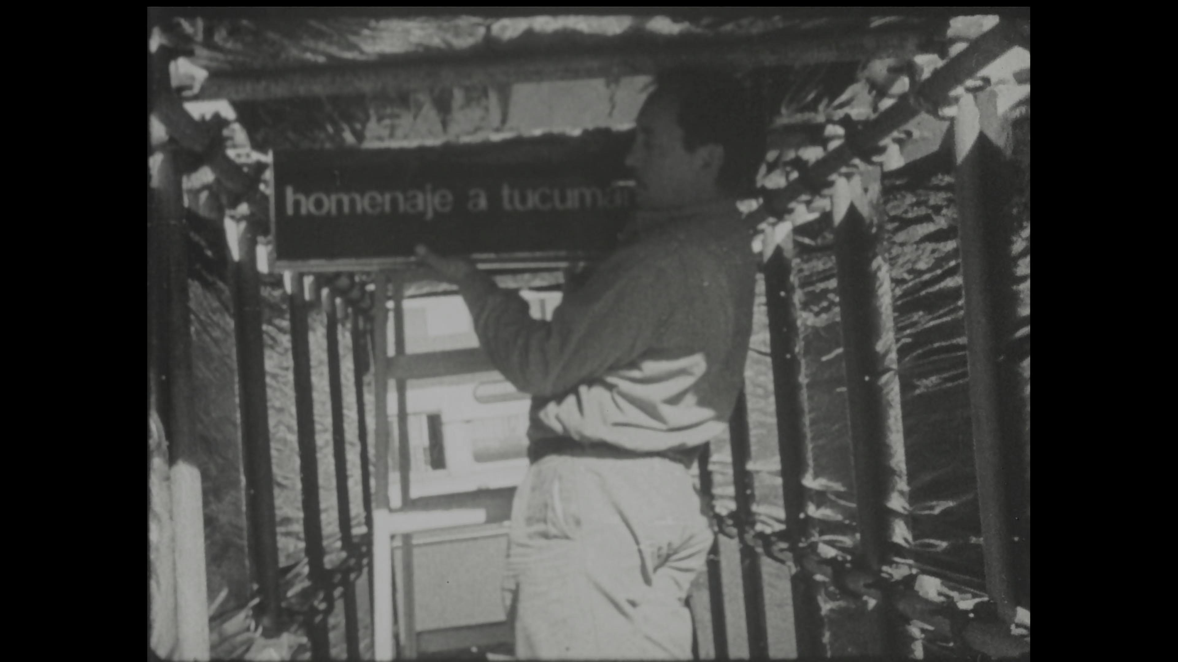 Black-and-white film still of a man on top of a ladder holding a large object while smoking