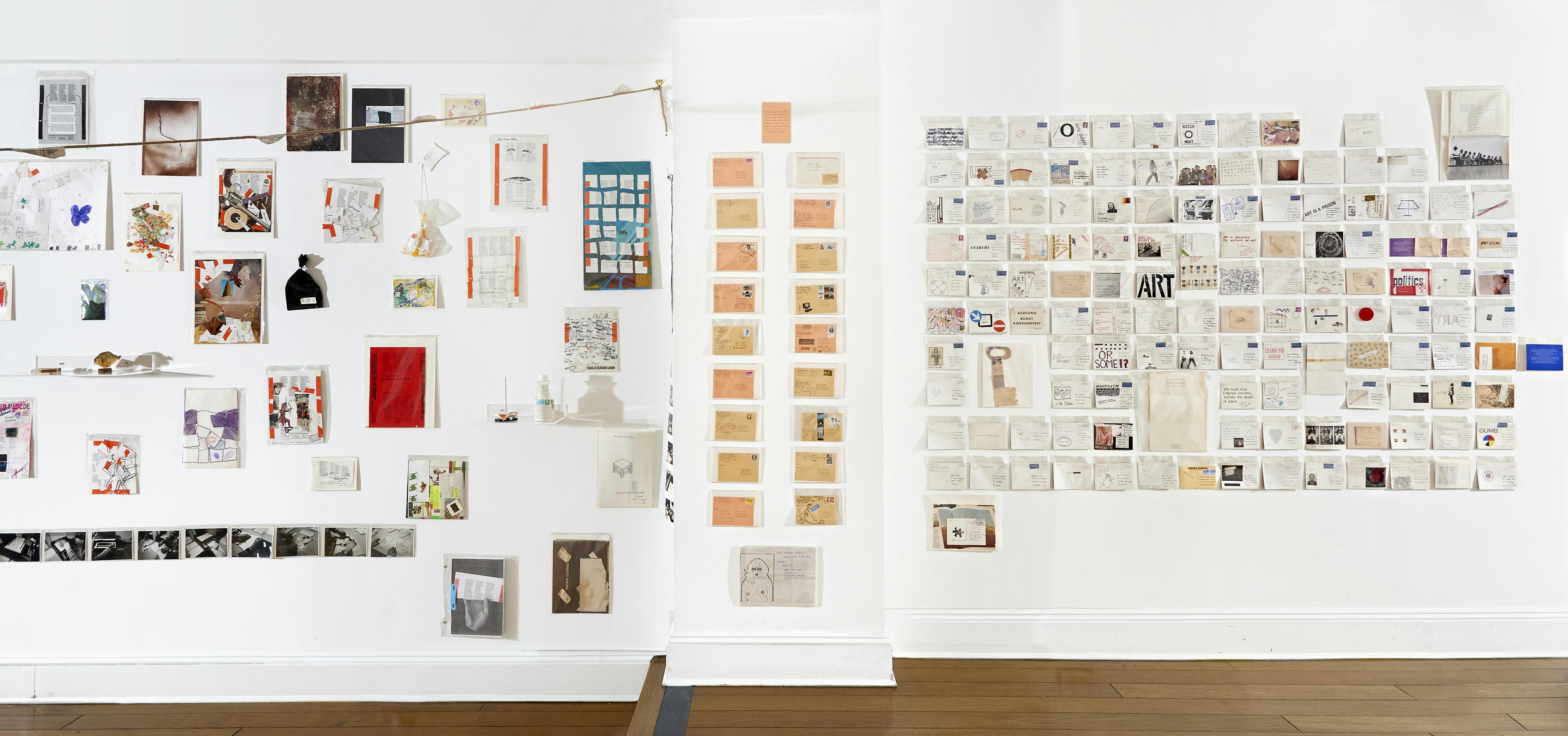 Installation view of Ulises Carrión exhibition showing small collages of varying size, photographs, and sketches mounted to a white wall.