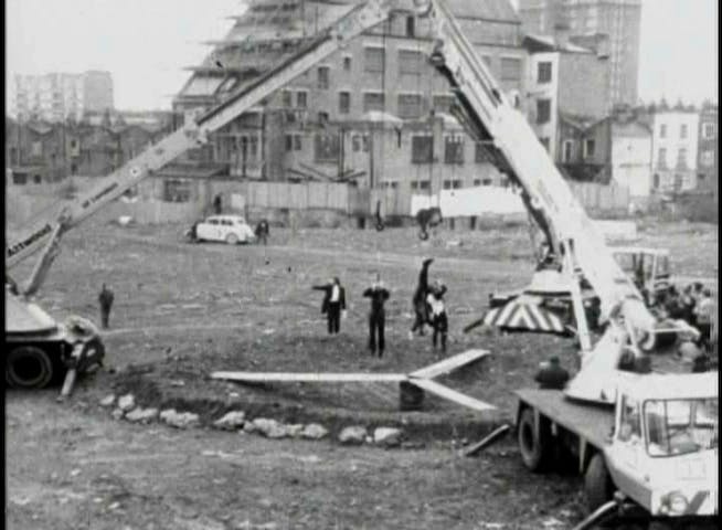 The image is a still from a black-and-white video documenting the performance of Leopoldo Maler’s Crane Ballet for the BBC. The arms of two mobile cranes extend from the image’s left and right edges to create a triangular shape. Below the apex of this triangle stands Maler, facing the camera and wearing a black tailcoat and white shirt in the style of an orchestral conductor. His right arm is extended toward the left edge of the picture. The cranes are parked on either side of a shallow pit, in the center of which three planks of wood extend outward at equal angles from the central point of an oil drum they are resting on.
