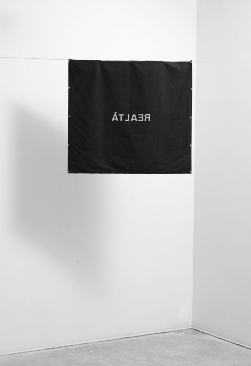 Black, square fabric hanging from a wire with the text "REALTÀ" written backwards in white it the center of the fabric.