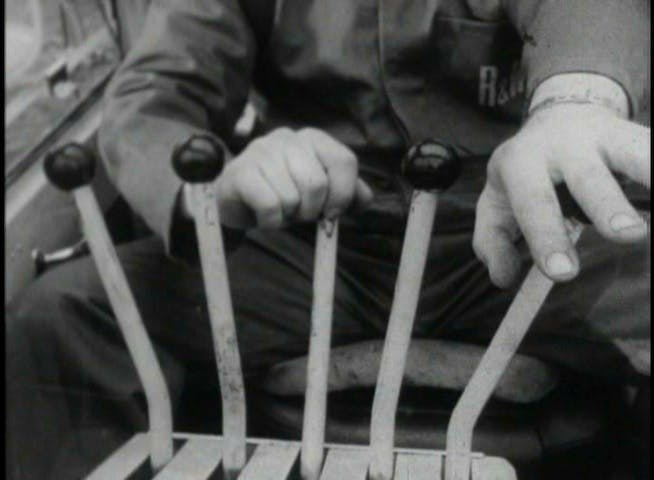 The image is a still from a black-and-white video documenting the performance of Leopoldo Maler’s Crane Ballet for the BBC. The image presents a close-up of the cab of a mobile crane, with five joysticks centered in the foreground. The crane driver, depicted from the shoulders down, has one hand on the middle joystick and the other hand outstretched toward the lever at the far right of the frame.