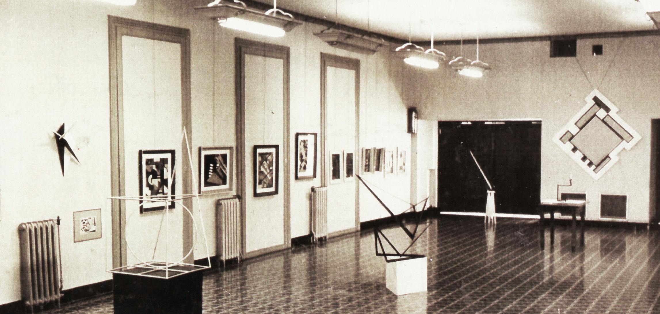 An installation view of paintings and four geometric sculptures on pedestals.