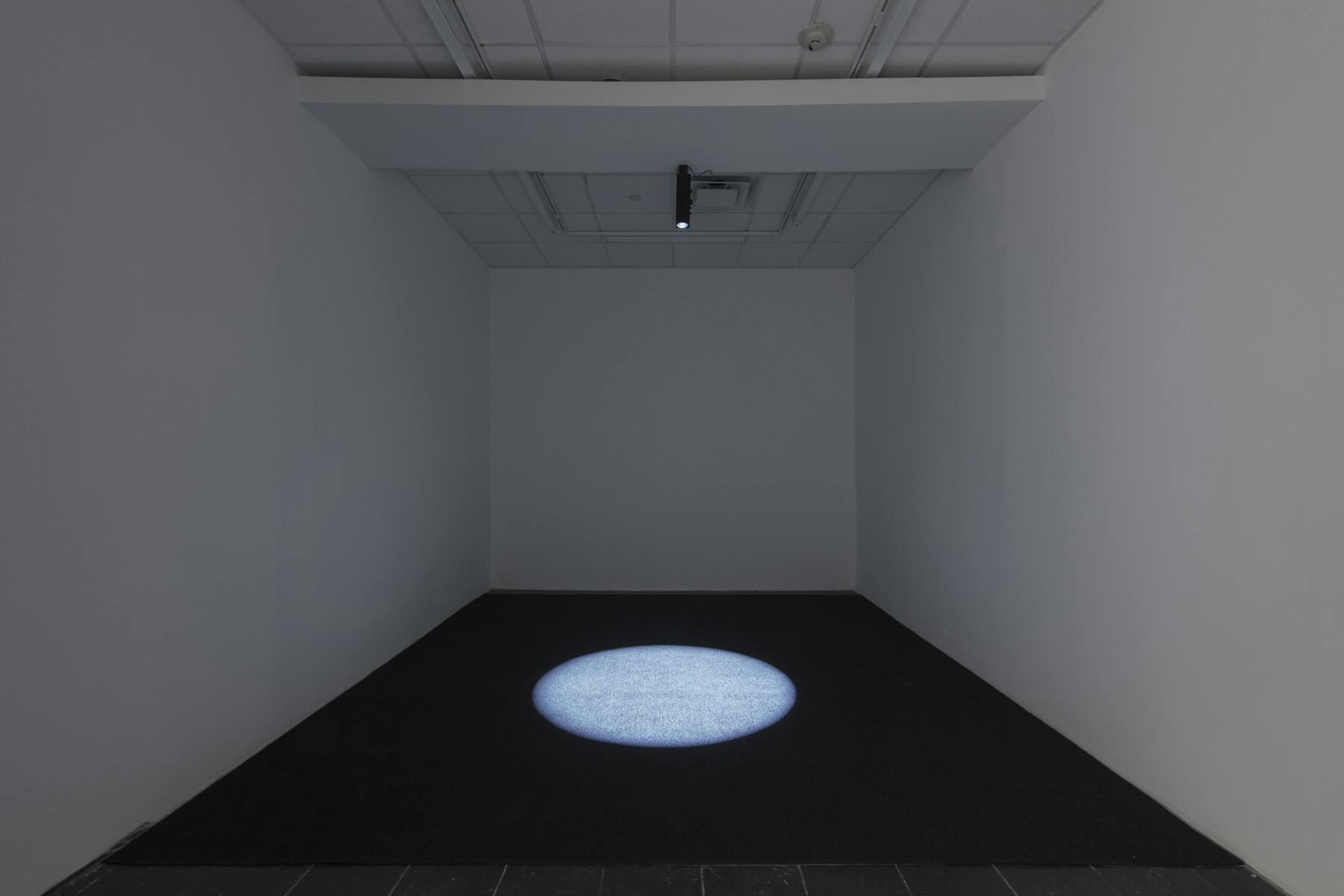 Installation view of a white-walled room with a ceiling-mounted light creating a bright circular shape on the floor.