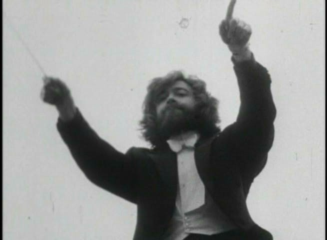 The image is a still from a black-and-white video documenting the performance of Leopoldo Maler’s Crane Ballet for the BBC. Maler fills the frame, gesturing with an orchestral baton in his right hand and with the index finger of his left hand extended. He wears a black tailcoat and white shirt in the style of an orchestral conductor.