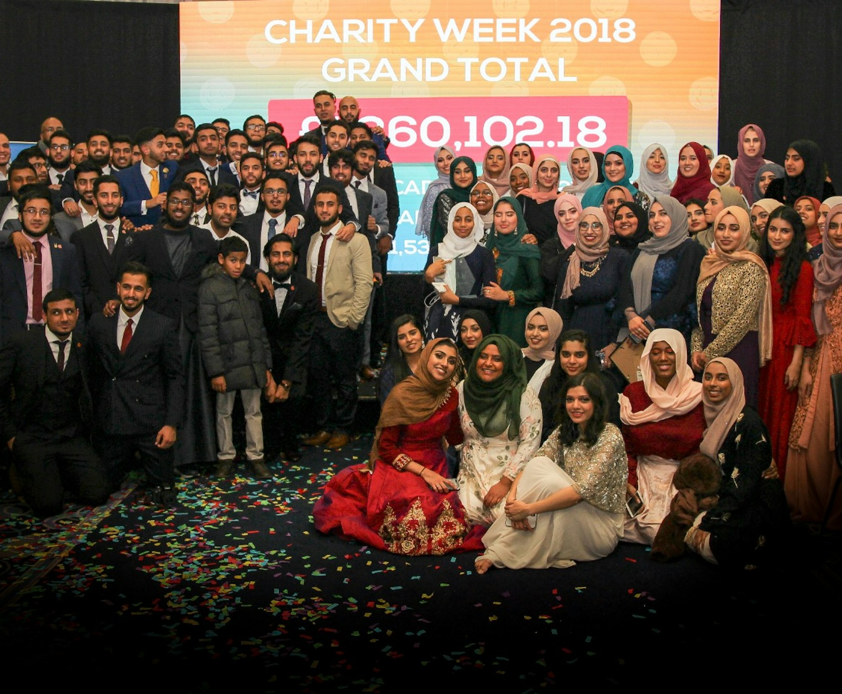 A large group gathers in front of a screen displaying 'Charity Week 2018 Grand Total: $960,102.18,' with confetti on the floor marking the celebration. Some background text is partially hidden by the crowd.