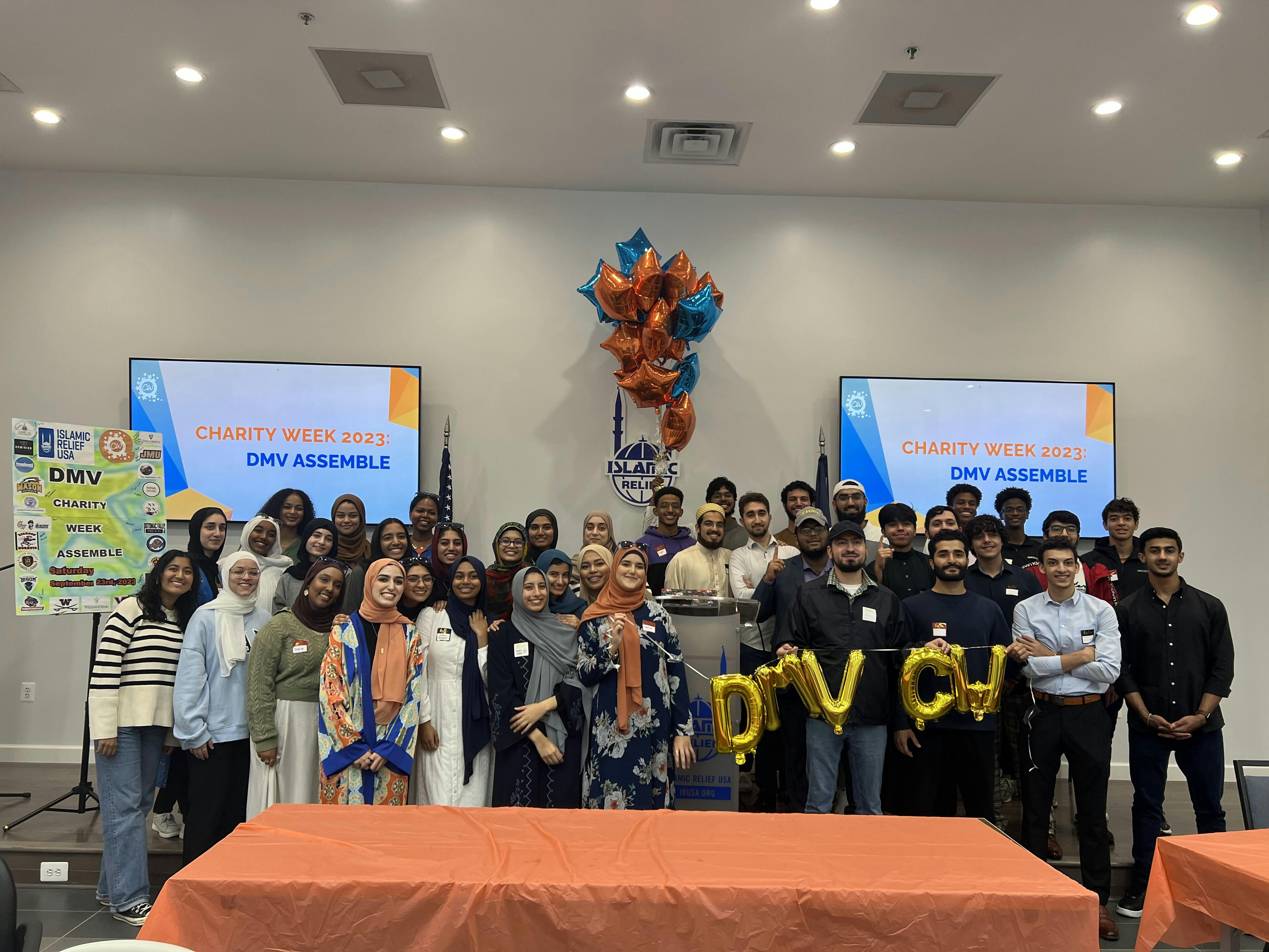 A group of attendees pose in front of orange-covered tables at a Charity Week 2023 event, with gold balloons spelling 'DMV CW' and screens behind them displaying 'DMV Assemble.' The setup includes a podium, festive balloons, and event signage.