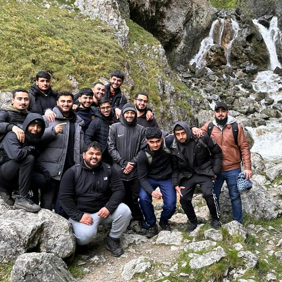 A group of individuals standing in front of a rocky hillside with a small waterfall in the background. They are dressed in warm clothing, indicating cool weather. The landscape features rugged terrain with rocks and sparse grass.
