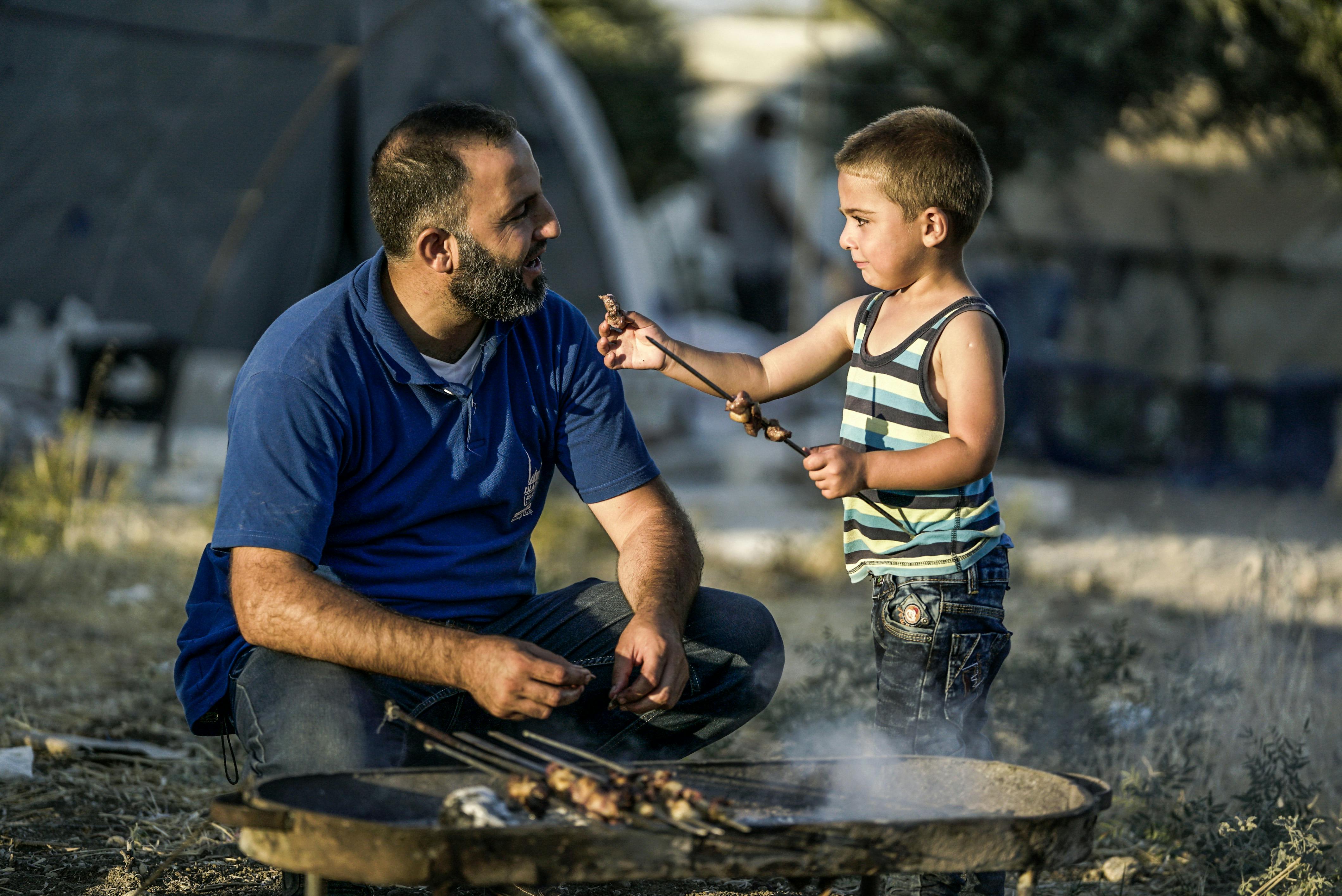 An adult kneels beside a round grill while a child offers a skewer of food, likely meat. They're outdoors near a tent, sharing a hands-on moment in a relaxed setting.
