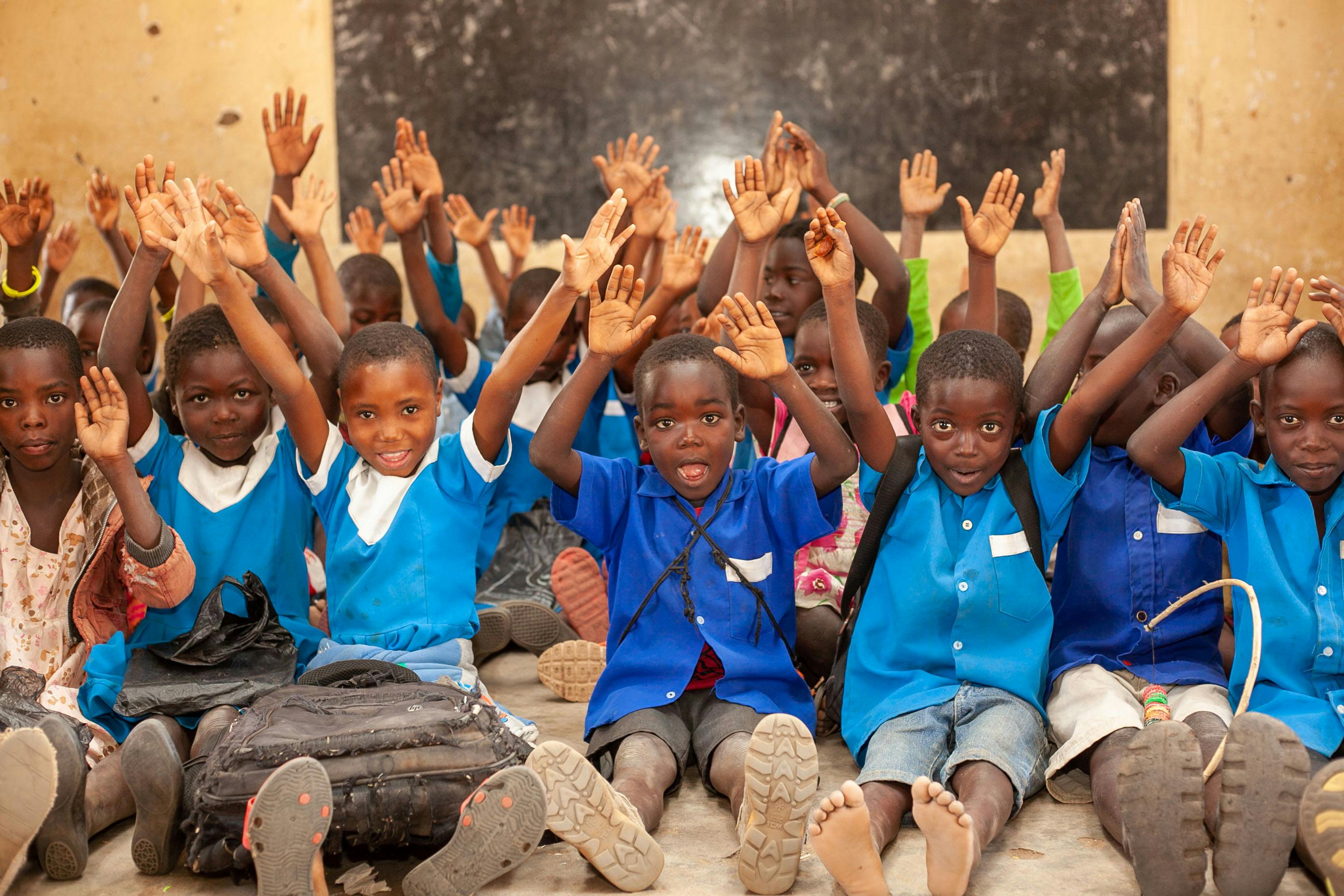 A group of children in blue uniforms sit on the ground with hands raised, likely participating in a classroom activity. A blackboard and backpacks in the background suggest a school setting.
