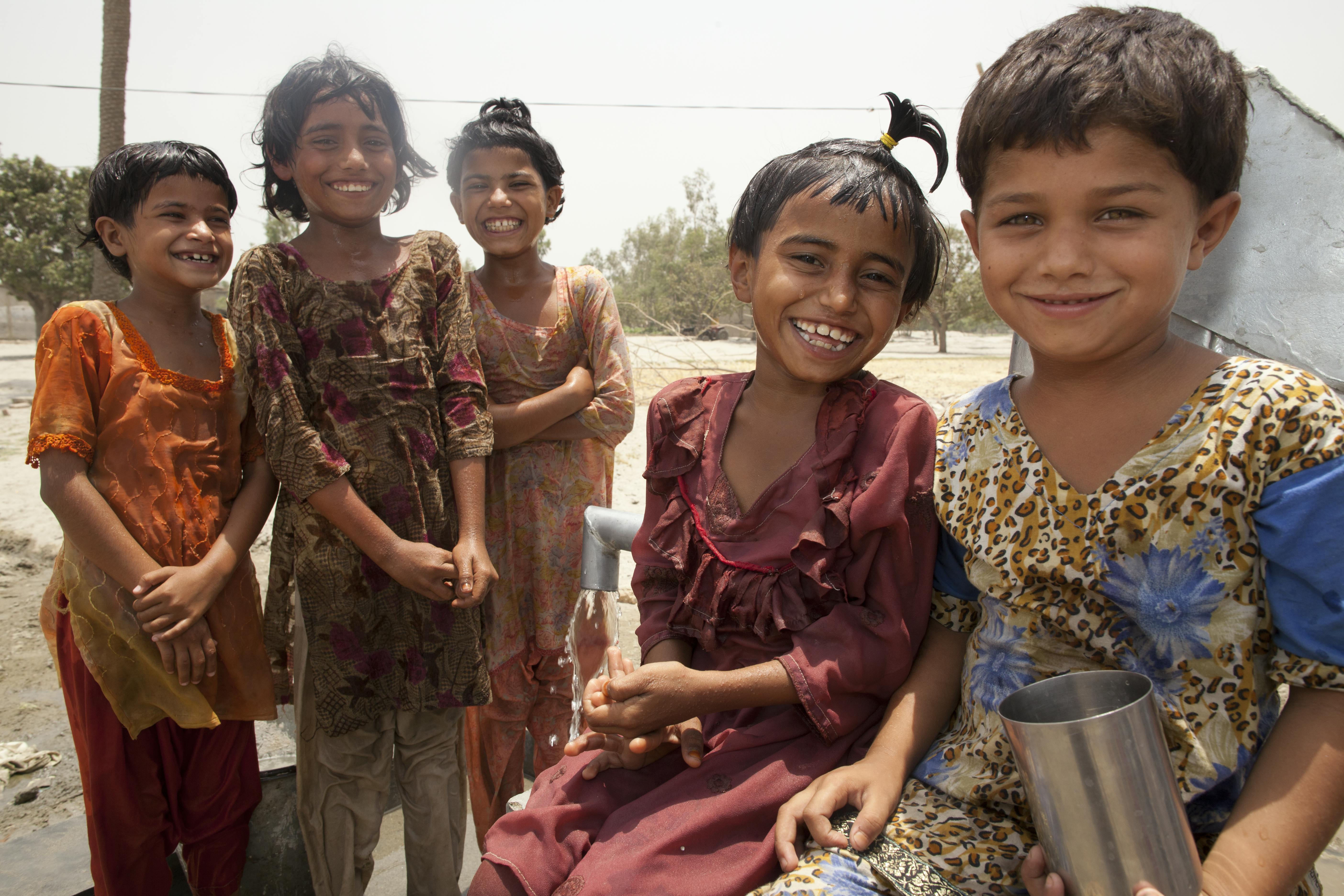  group of children dressed in traditional clothing are gathered outdoors in a rural area. Some are standing, others sitting, and one child is holding a metal cup. The background includes trees and a dirt road.