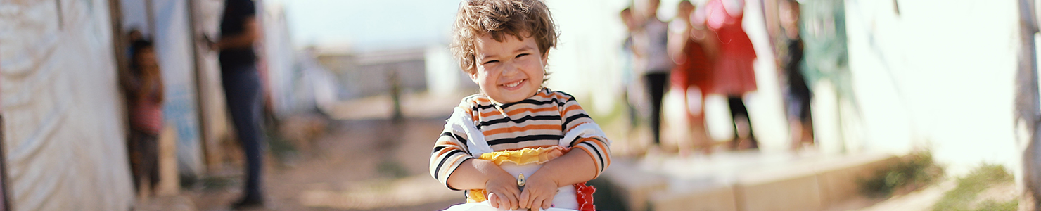 A young child in a striped shirt holds a white plastic bag while standing in an outdoor area that resembles a temporary settlement. Several people are visible in the background along a narrow pathway between structures.