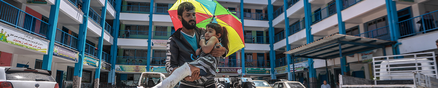 An individual holds a child under a vibrant umbrella outside a multi-story building with blue railings and Arabic banners. Parked cars line the front, adding to the everyday street scene.