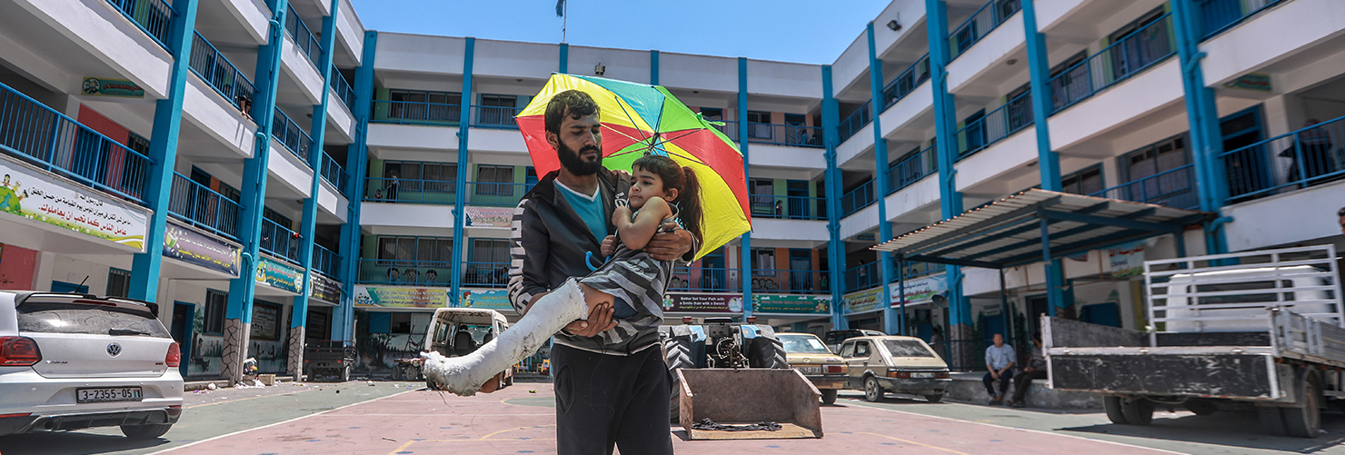 An individual holds a child under a vibrant umbrella outside a multi-story building with blue railings and Arabic banners. Parked cars line the front, adding to the everyday street scene.