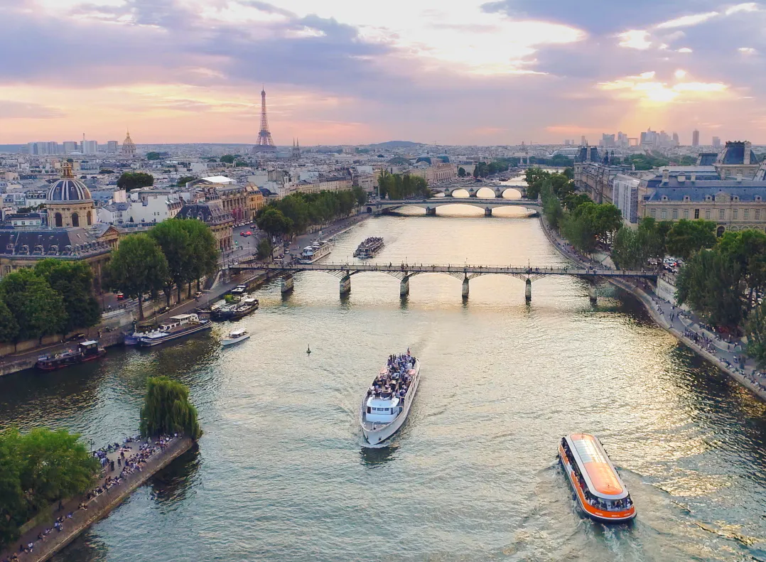 Mynd af bátum á ánni Signu með hús og tré Parísar í bakgrunni
