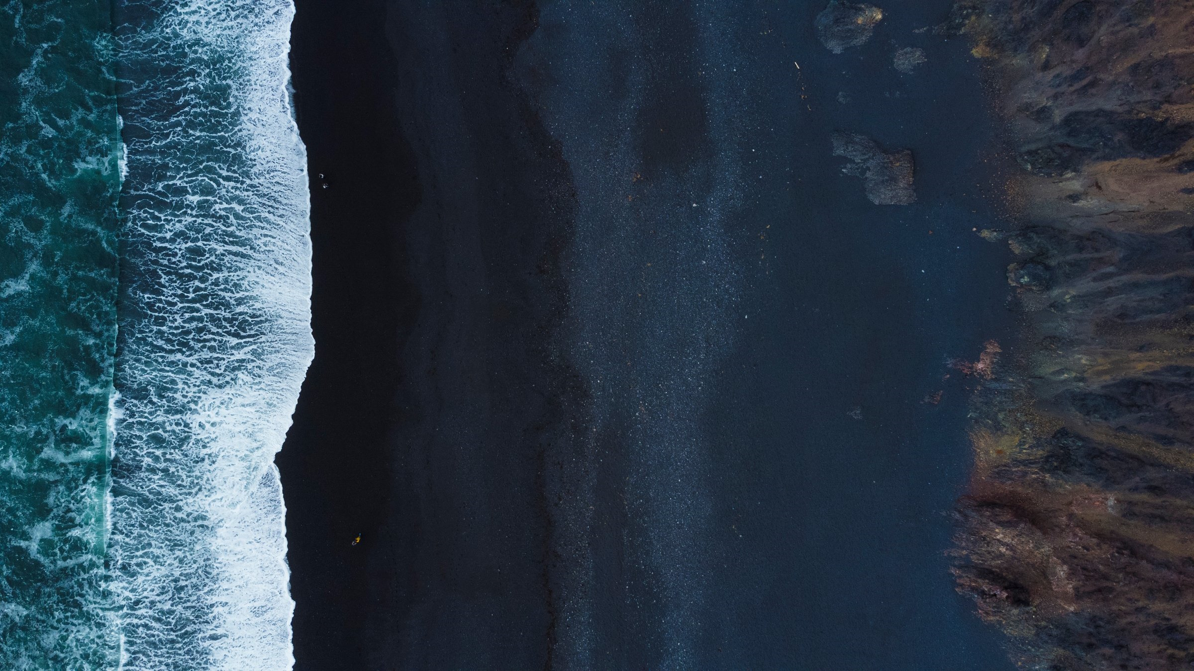 Loftmynd af svartri sandströnd þar sem hvítar öldur brotna að landi. Dökkt fjörusandur og grunnsævi mynda sterkt mynstur frá sjó að landi, með nokkrum klettum sýnilegum í sjónum.