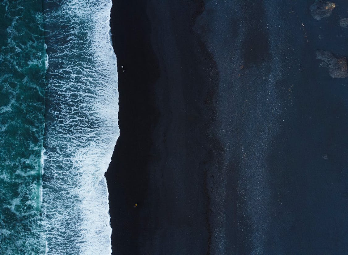 Loftmynd af svartri sandströnd þar sem hvítar öldur brotna að landi. Dökkt fjörusandur og grunnsævi mynda sterkt mynstur frá sjó að landi, með nokkrum klettum sýnilegum í sjónum.
