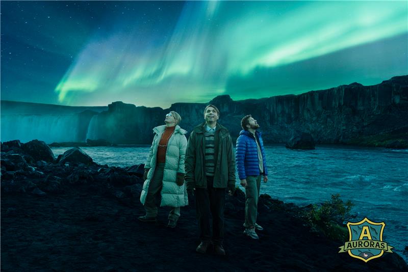 Three people stand beneath vibrant northern lights along a rugged Icelandic coastline, featured in Business Iceland’s AURORAS campaign promoting winter travel and natural wonders.