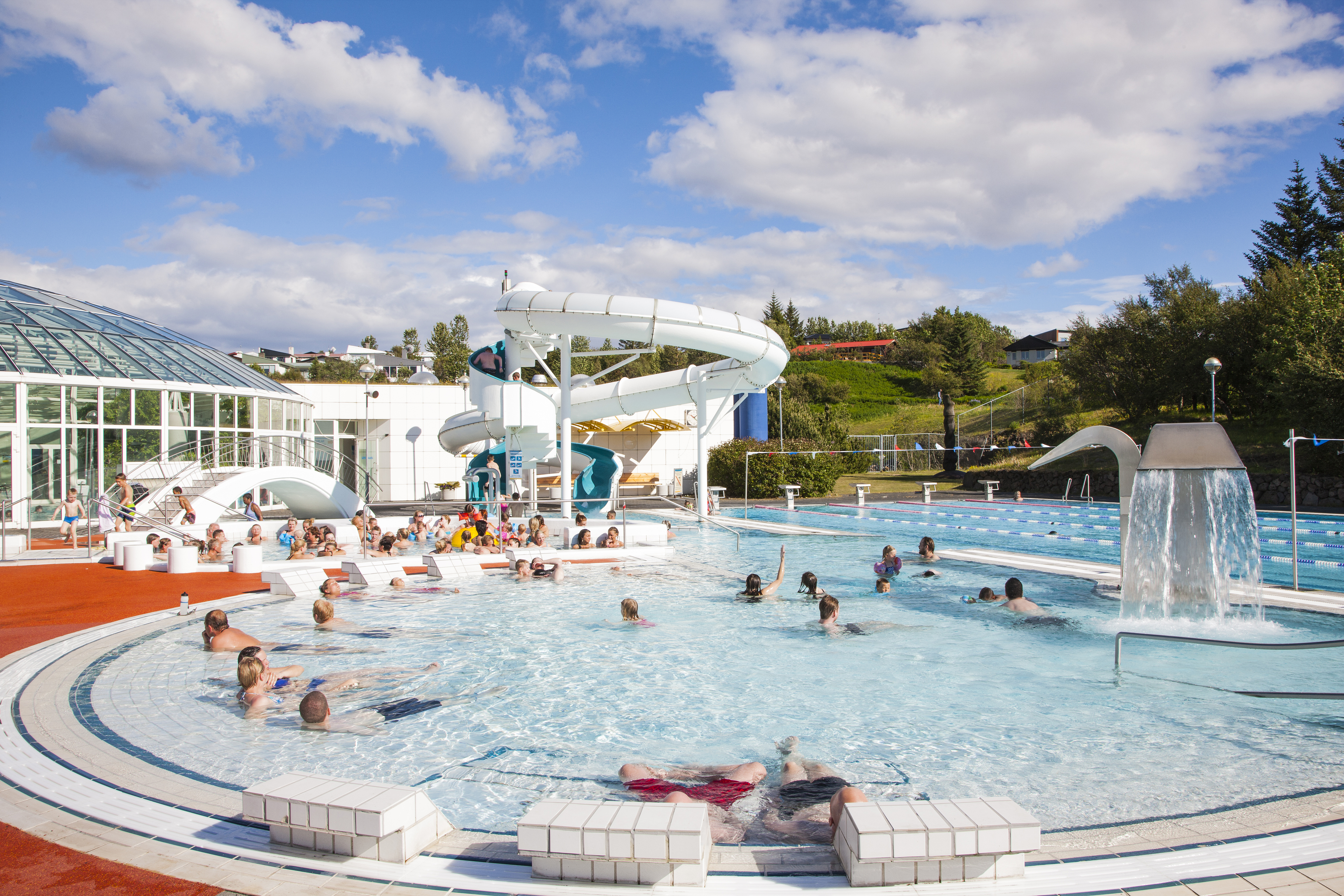 Fólk nýtur sín í útisundlaug Árbæjarlaugar á sólskinsdegi, sumir í pottum en aðrir að synda eða leika sér við vatnsrennibraut.