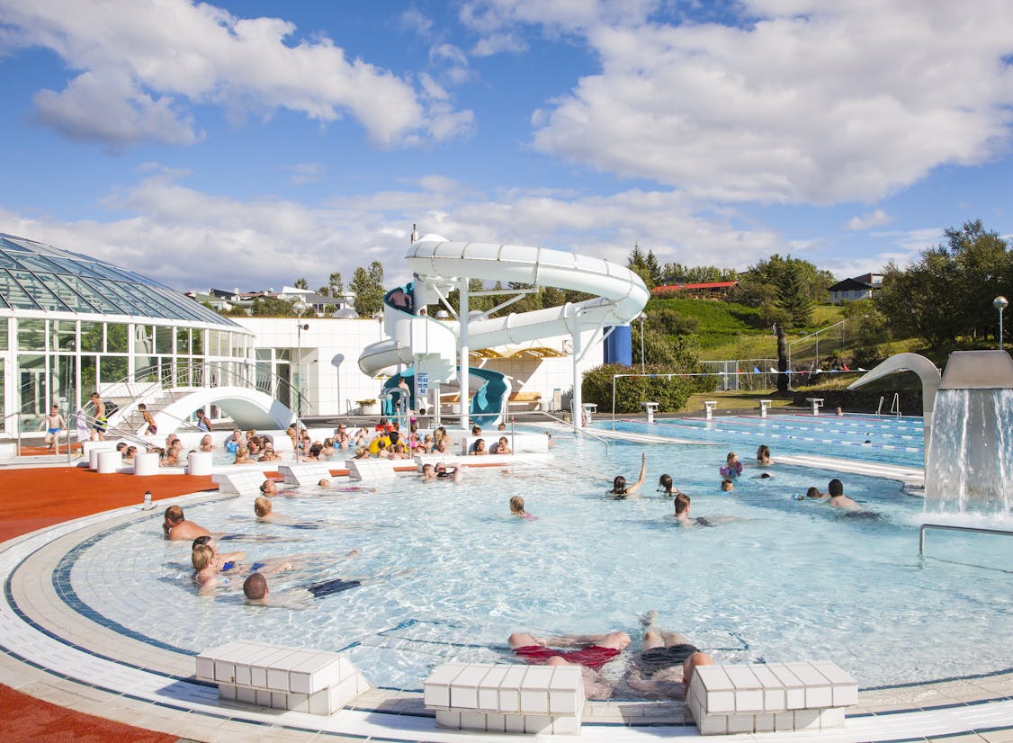 Fólk nýtur sín í útisundlaug Árbæjarlaugar á sólskinsdegi, sumir í pottum en aðrir að synda eða leika sér við vatnsrennibraut.