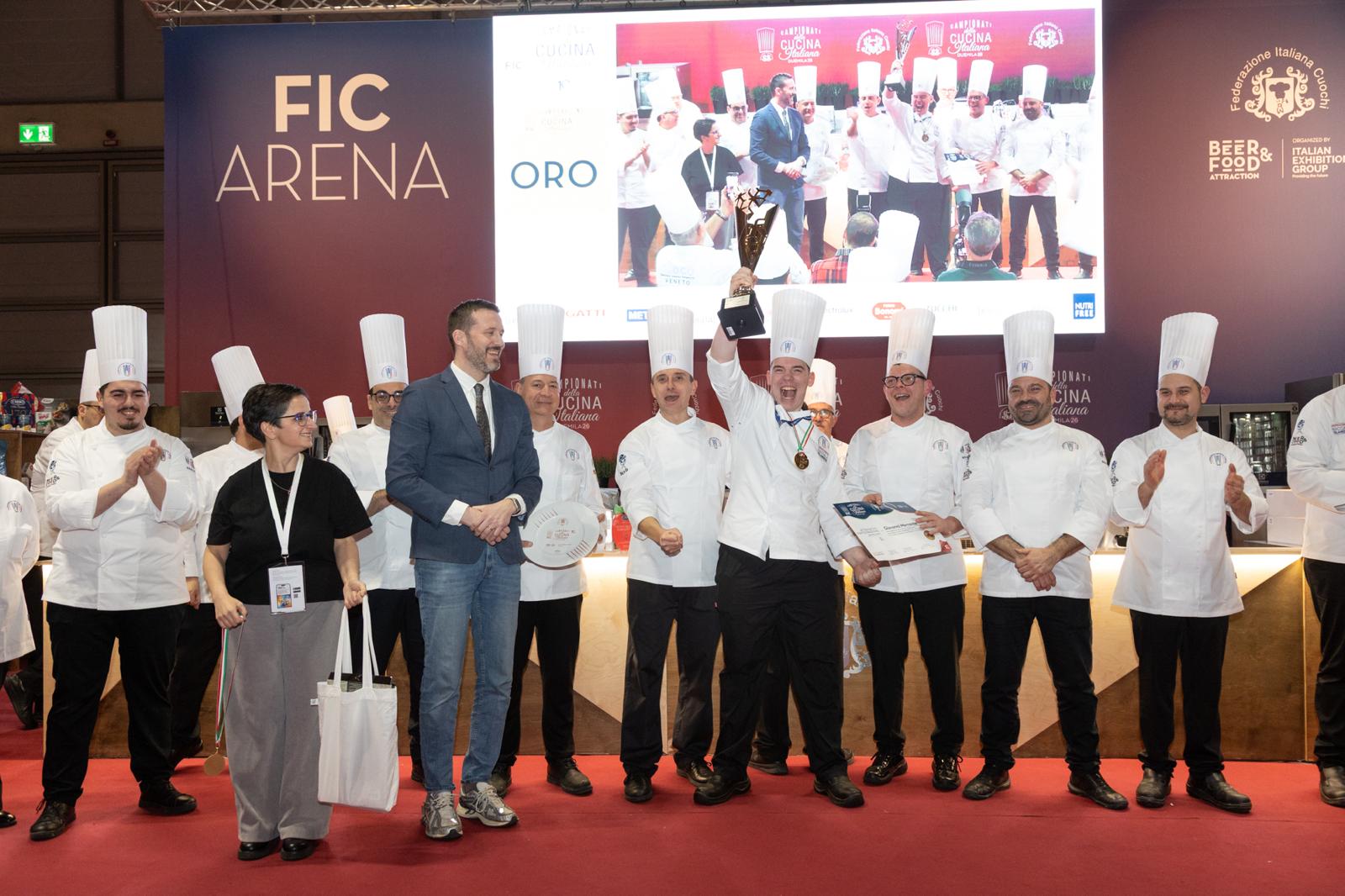 Kokkaskólakeppni á matvælasýningu í Rimini þar sem hópur kokka í hvítum einkennisbúningum og háum kokkahúfum stendur á sviði. Sigurvegari heldur verðlaunagrip á lofti og fagnar, á meðan aðrir keppendur og viðstaddir klappa. Keppnin var skipulögð af Bacalao de Islandia