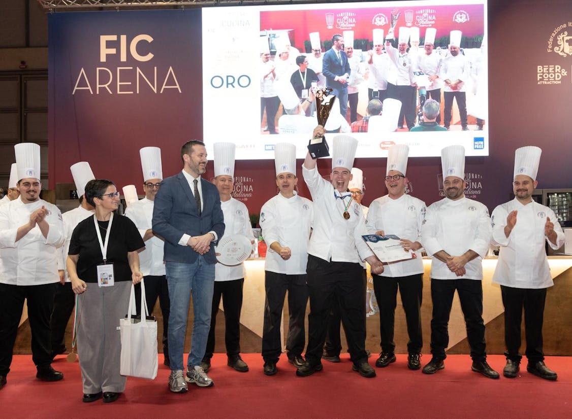 Kokkaskólakeppni á matvælasýningu í Rimini þar sem hópur kokka í hvítum einkennisbúningum og háum kokkahúfum stendur á sviði. Sigurvegari heldur verðlaunagrip á lofti og fagnar, á meðan aðrir keppendur og viðstaddir klappa. Keppnin var skipulögð af Bacalao de Islandia