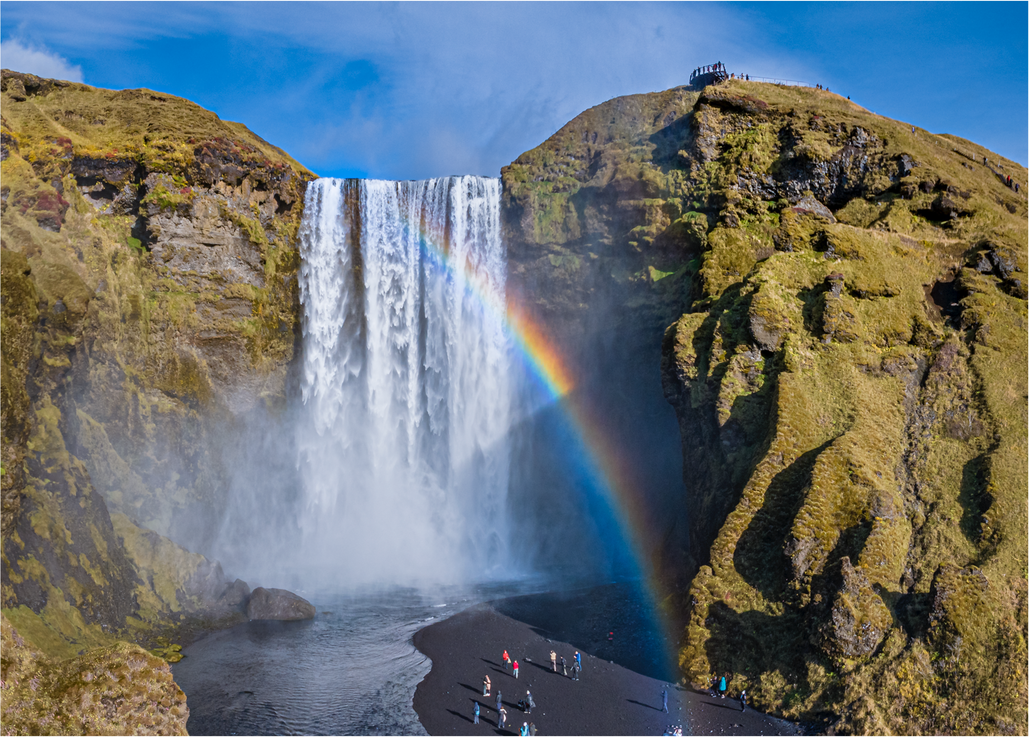 Skógafoss fellur fram af háum klettum á Suðurlandi, með regnboga í úðanum og gesti sem ganga á svörtum sandinum við fossinn.