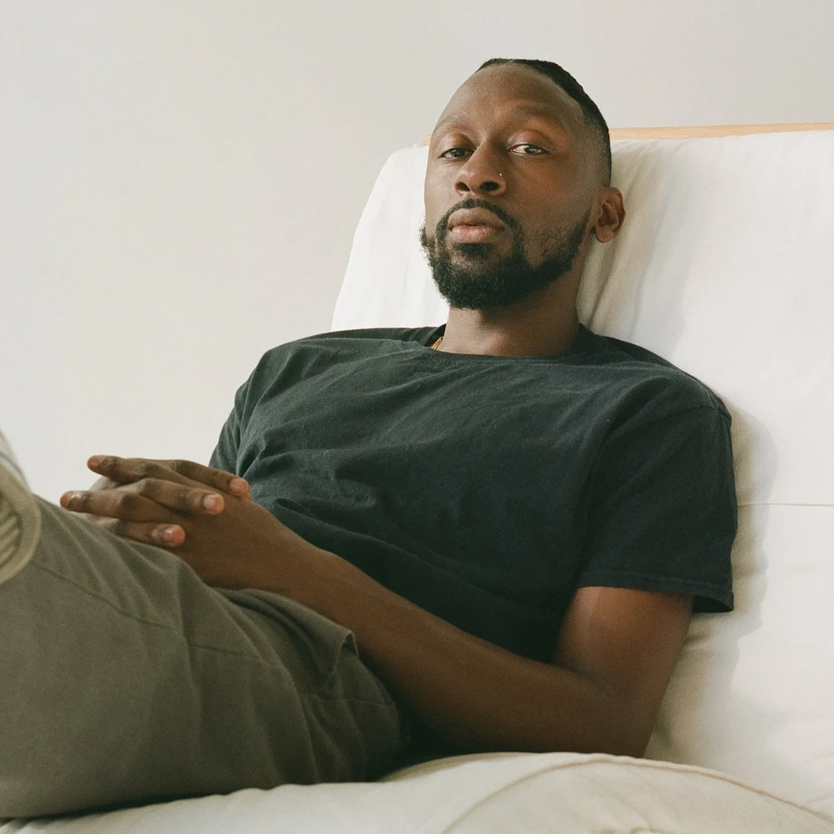 Brandon Thomas Brown reclines on a white sofa in a soft gray t-shirt, hands resting in his lap, gaze direct and composed against a neutral wall.