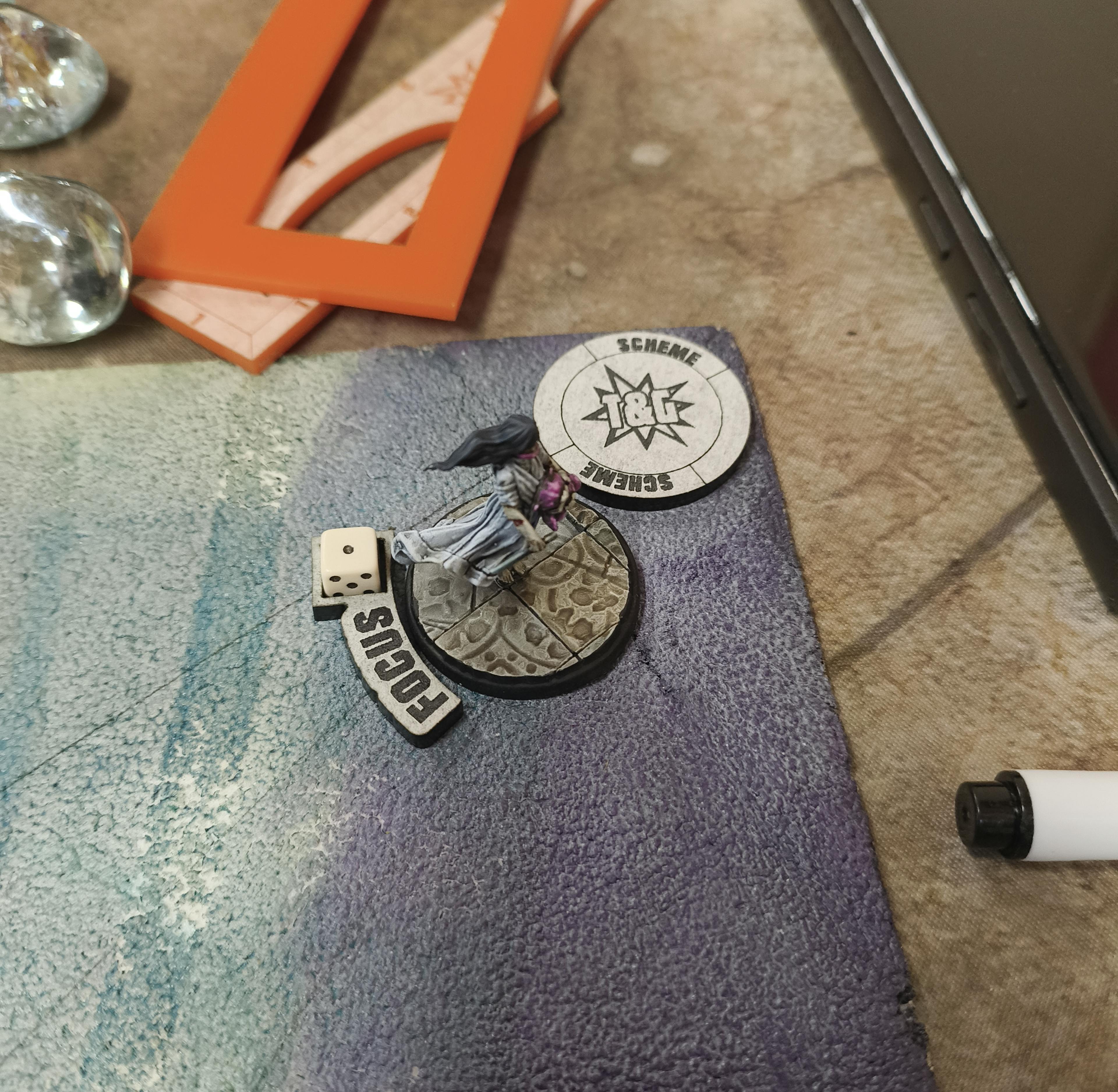A Focused Crooligan next to a Scheme Marker in the corner of a Malifaux board.