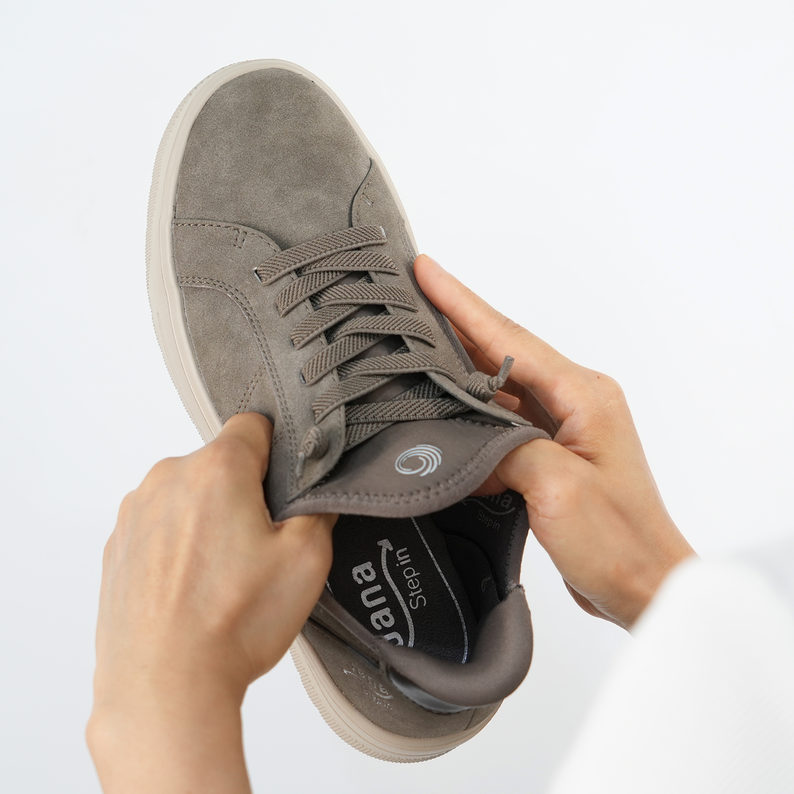 Person zieht den Schaft eines grau-braunen Jana Step in Sneakers auseinander – dies zeigt wie elastisch die Senkel und der Schaft sind, was das reinschlüpfen möglich macht.