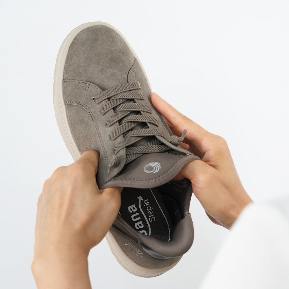 Person zieht den Schaft eines grau-braunen Jana Step in Sneakers auseinander – dies zeigt wie elastisch die Senkel und der Schaft sind, was das reinschlüpfen möglich macht.