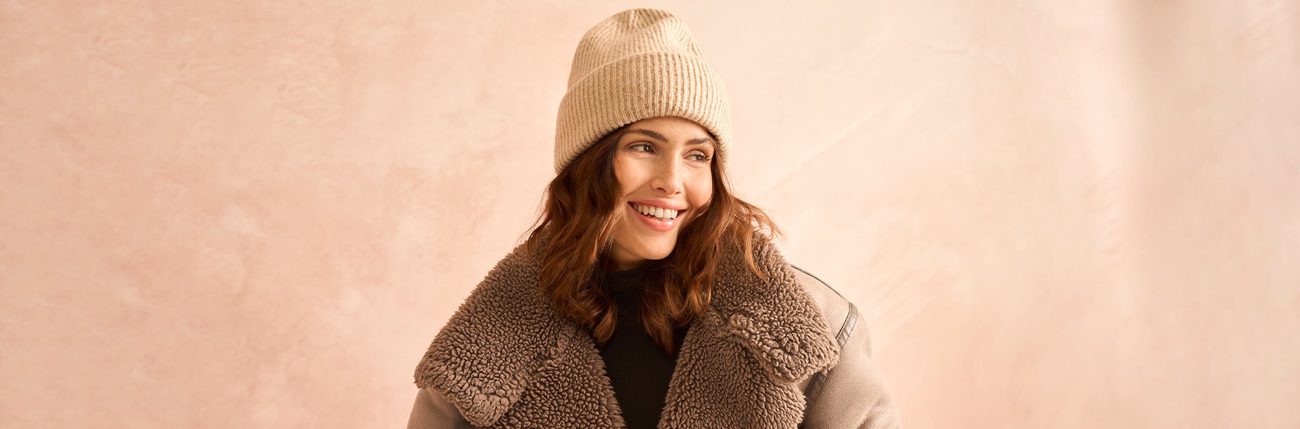 Model trägt eine braune Winterjacke mit Fellkragen und eine beige Mütze