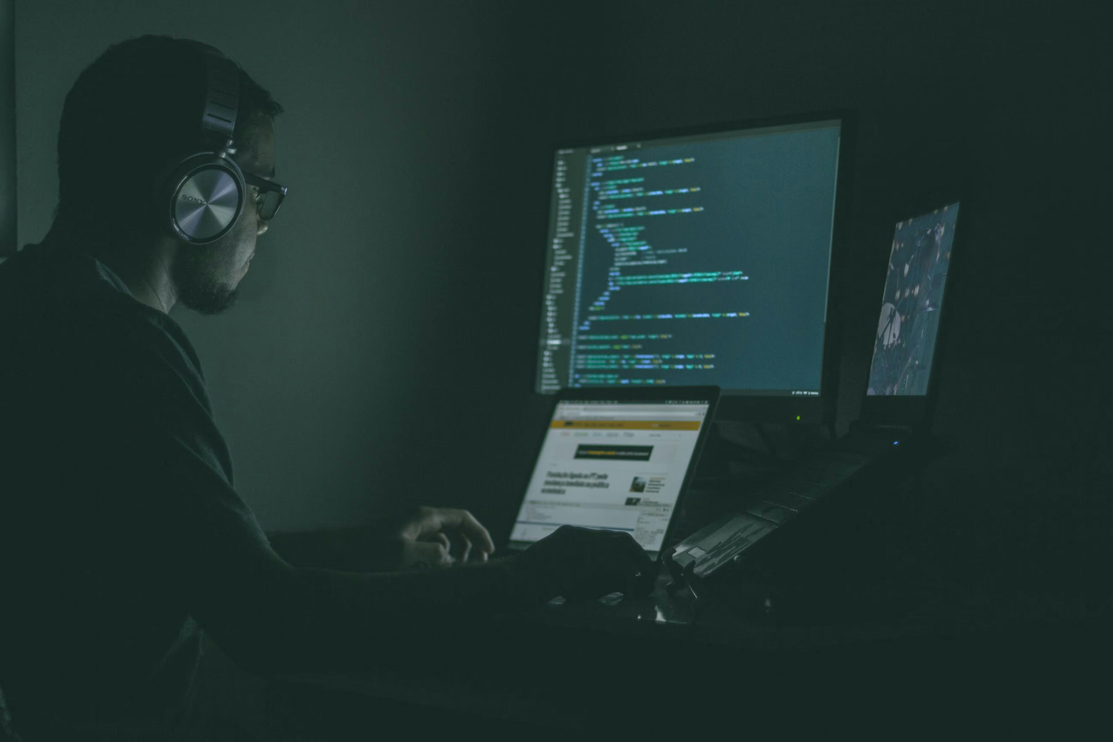 Guy at desk coding on multiple screens in the dark with headphones on