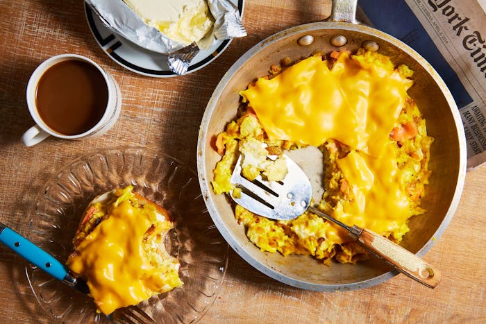 Eggs with lox and onions topped with melted cheese alongside coffee, butter and a New York Times newspaper atop a wooden surface.