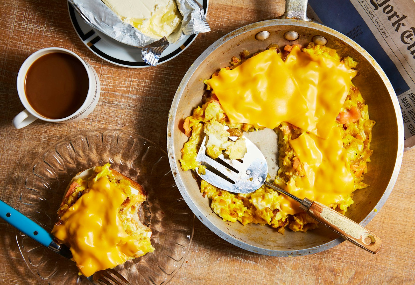 Eggs with lox and onions topped with melted cheese alongside coffee, butter and a New York Times newspaper atop a wooden surface.