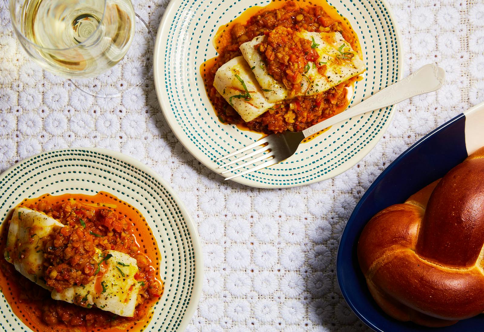 Two servings on Moroccan fish with chickpea stew alongside white wine and loaf of challah, atop white tablecloth.