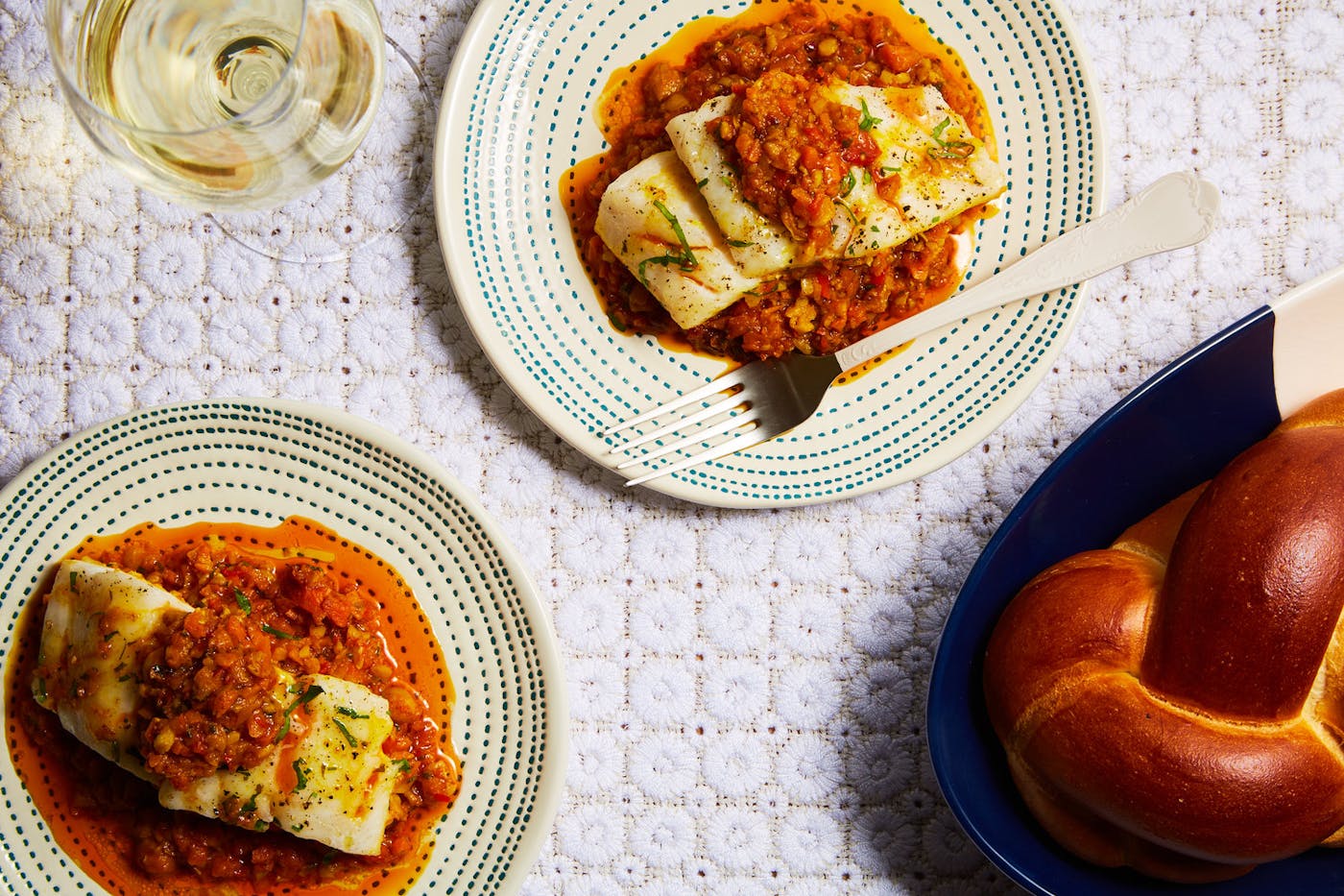 Two servings on Moroccan fish with chickpea stew alongside white wine and loaf of challah, atop white tablecloth.