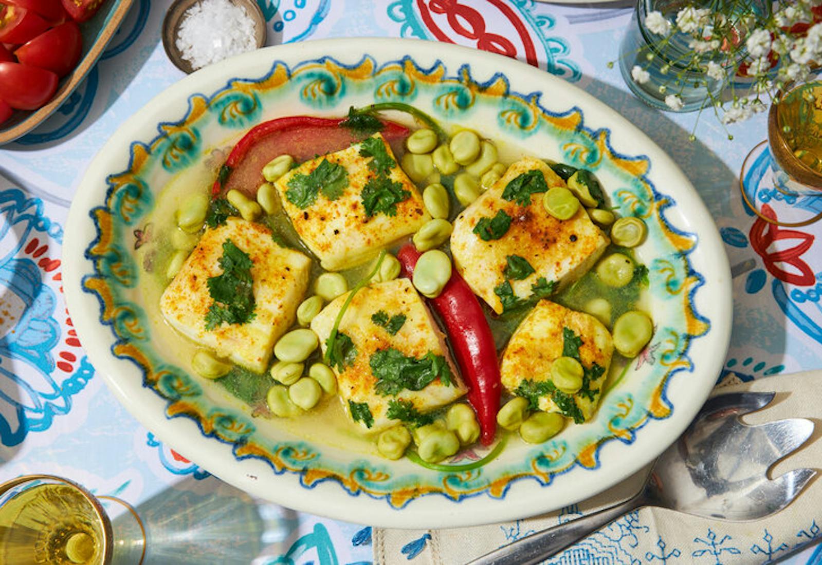 Pescado picante garnished with cilantro, bowl of sliced cherry tomatoes, white wine, fresh flowers atop vibrant tablecloth.