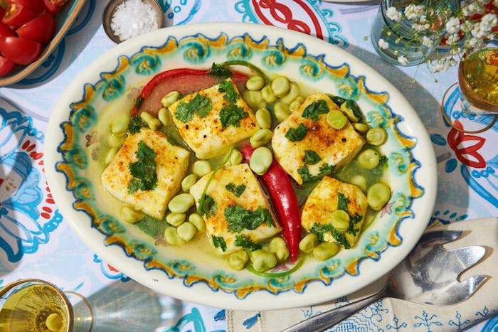 Pescado picante garnished with cilantro, bowl of sliced cherry tomatoes, white wine, fresh flowers atop vibrant tablecloth.