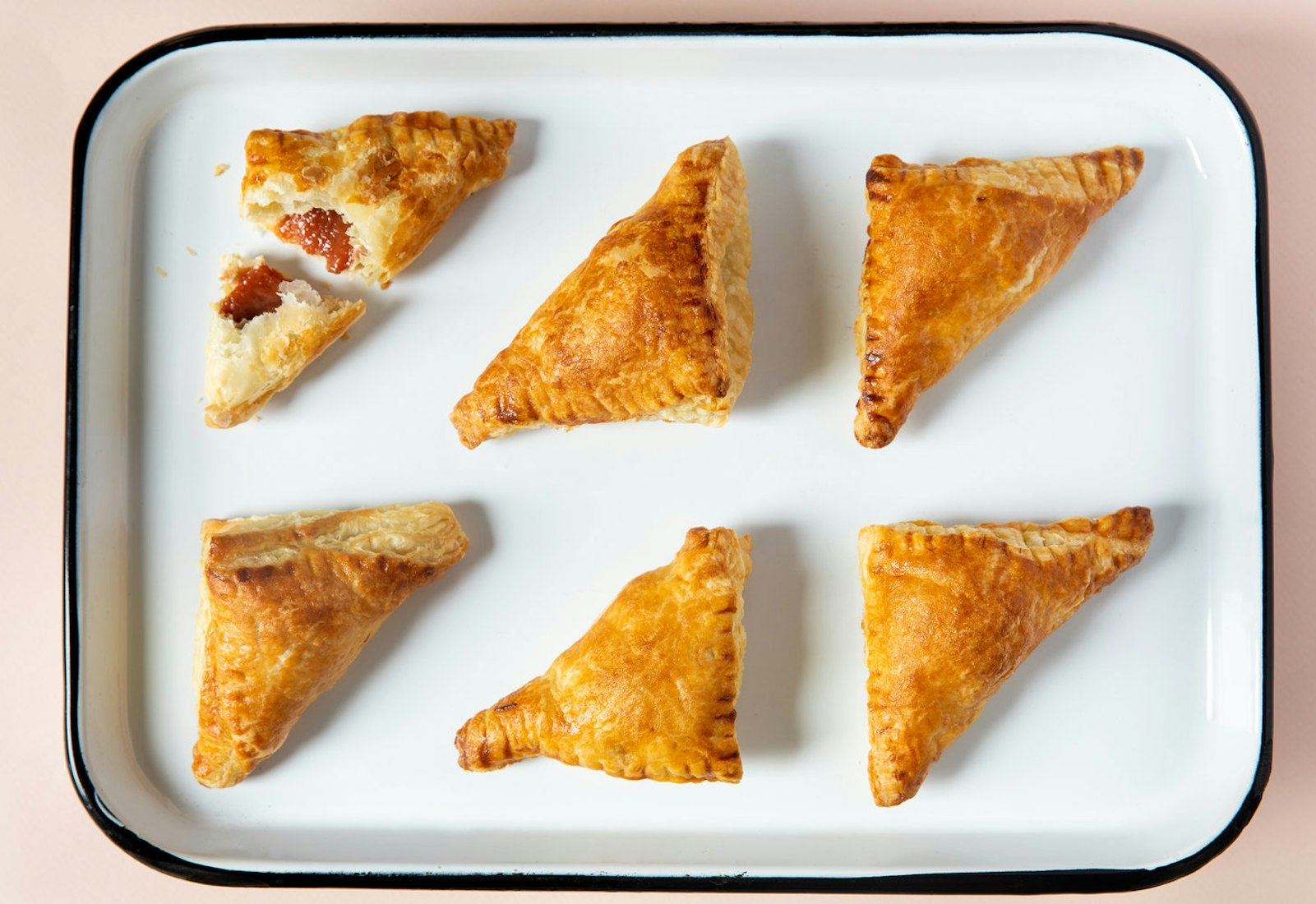 Golden pastelitos on white baking pan atop pale pink surface.