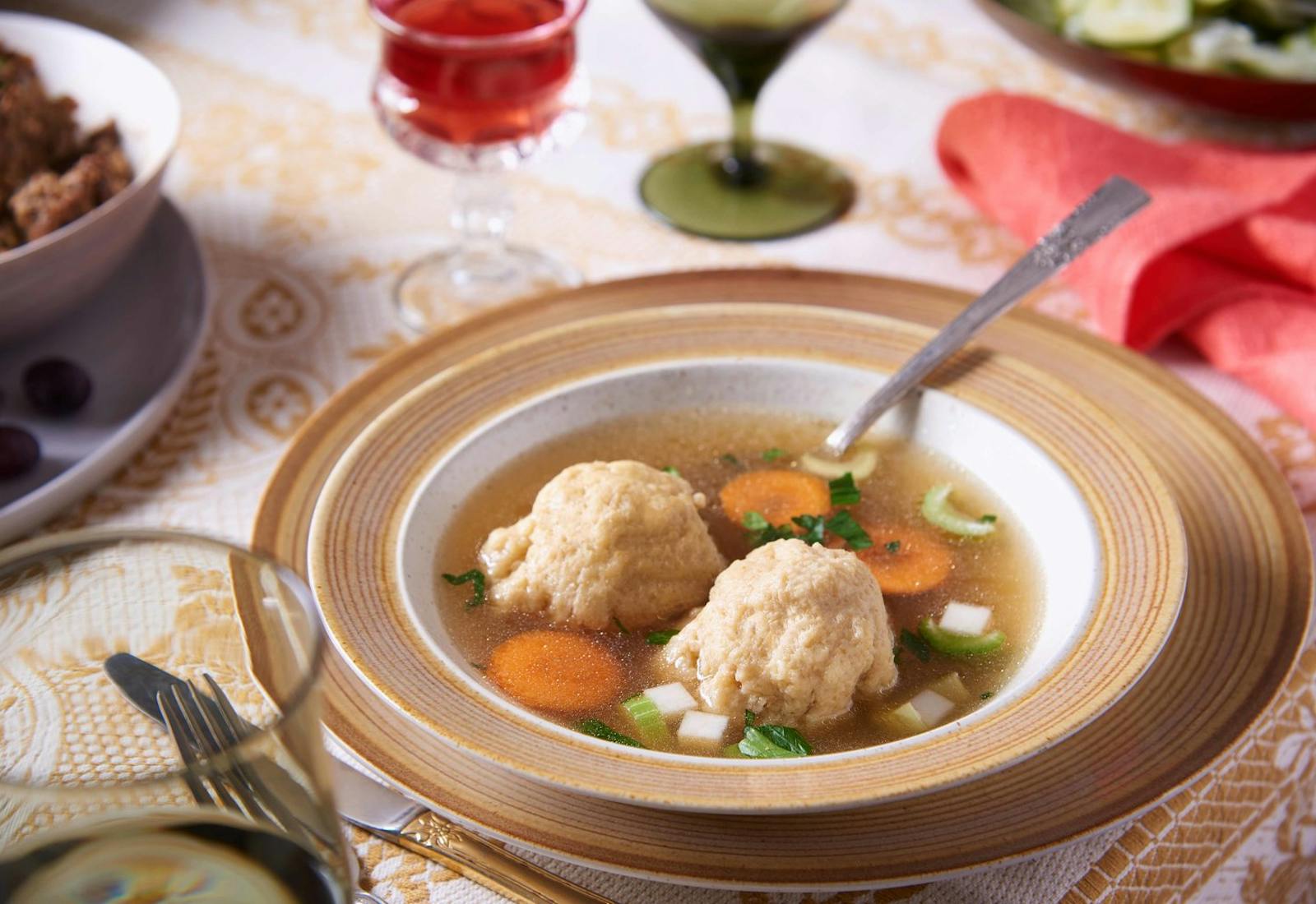 Serving of matzo ball soup in brown-rimmed bowl atop matching plate, alongside glasses of wine and pink napkin.