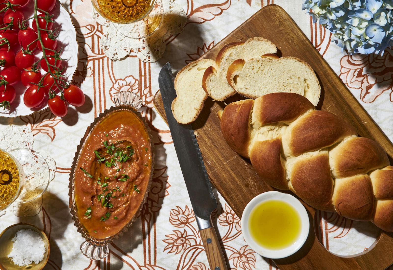 Kyopolou with chopped parsley in glass dish alongside sliced water challah, cherry tomatoes on the vine, and glasses of white wine.