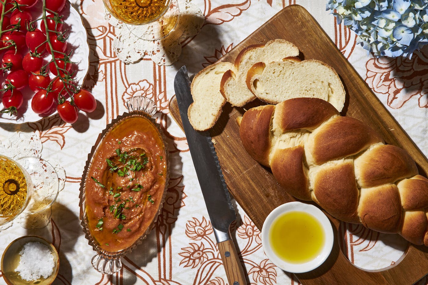 Kyopolou with chopped parsley in glass dish alongside sliced water challah, cherry tomatoes on the vine, and glasses of white wine.